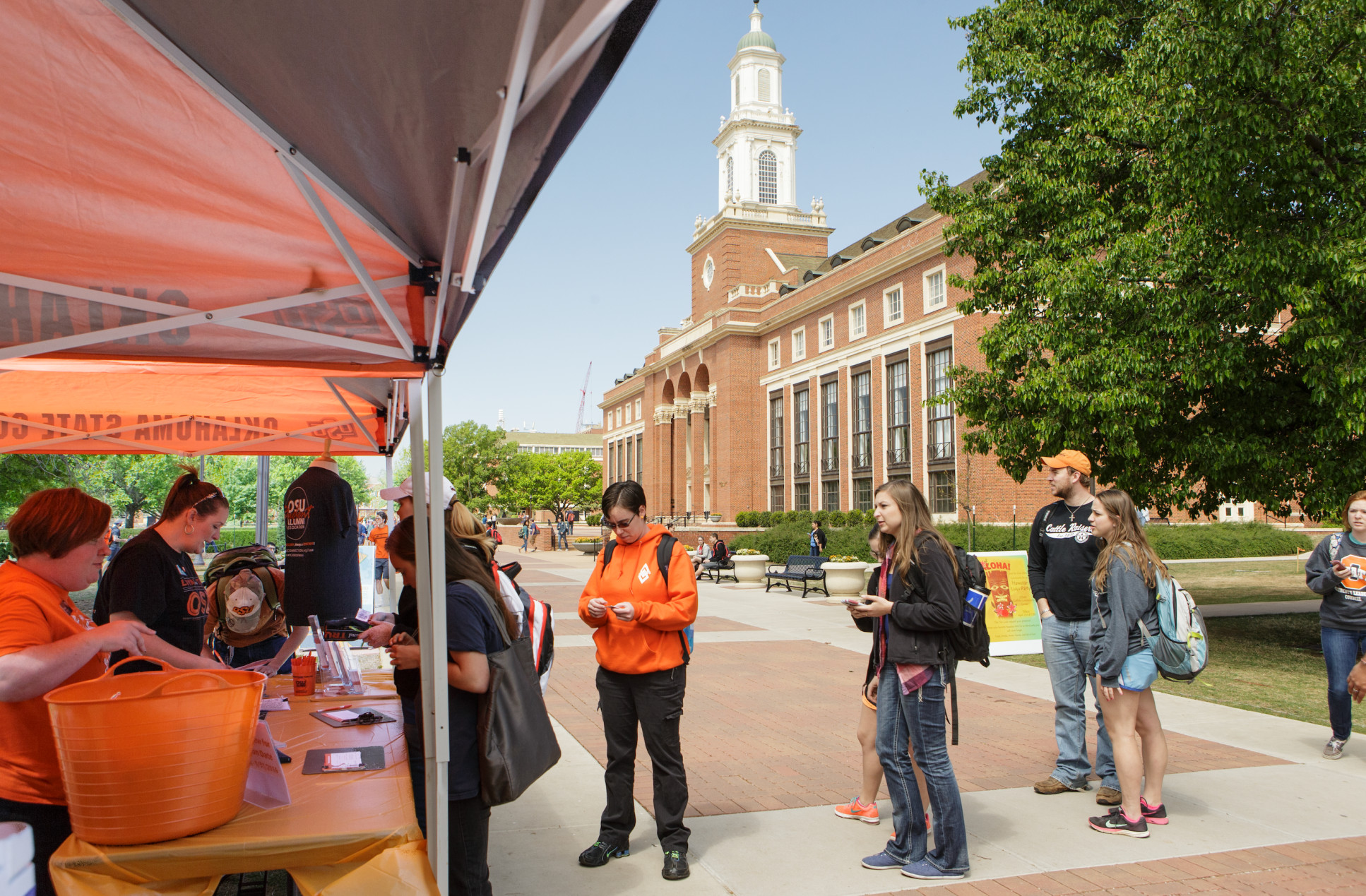Group of people gathered outdoors on a college campus near a tent, with some interacting with staff or volunteers. A large brick building with white columns and a clock tower is visible in the background. Others are standing, using their phones, or chatting. Clear daytime weather.