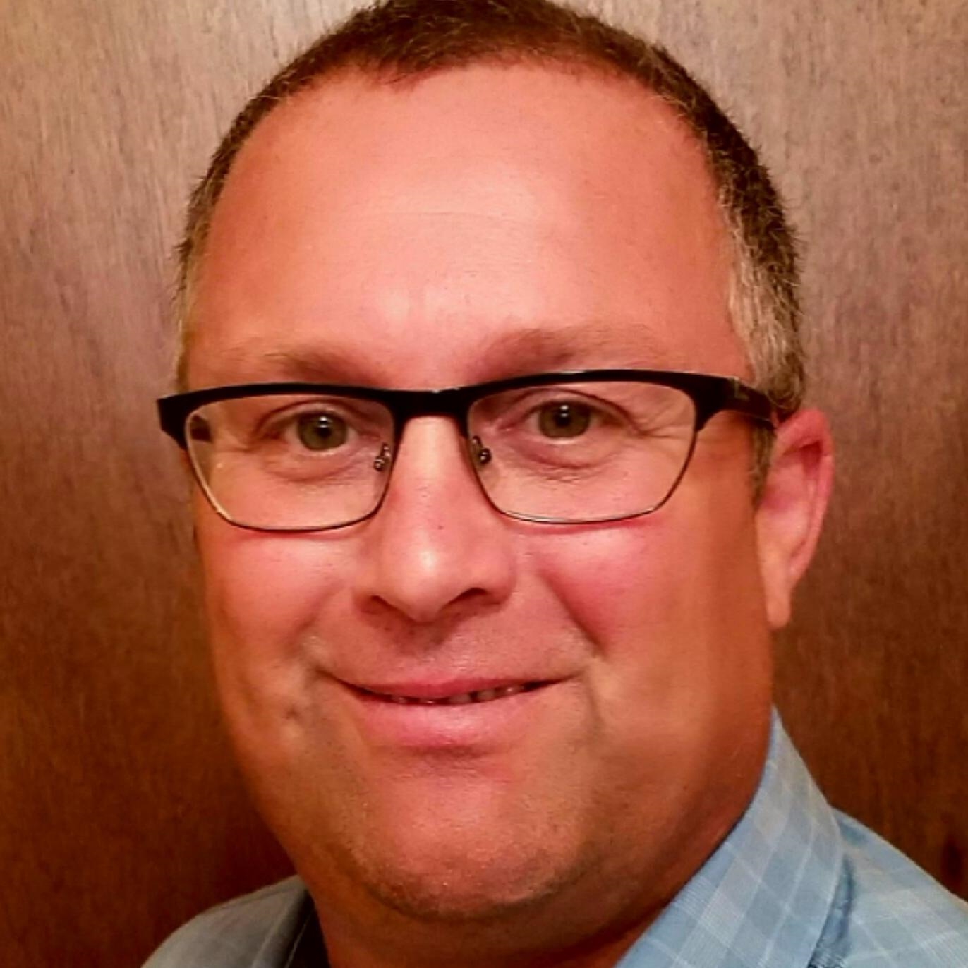 A headshot of a man with short hair and glasses wearing a blue check shirt, standing in front of a brown background.