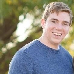 A man with brown hair and a blue shirt smiles with a natural background of greenery.