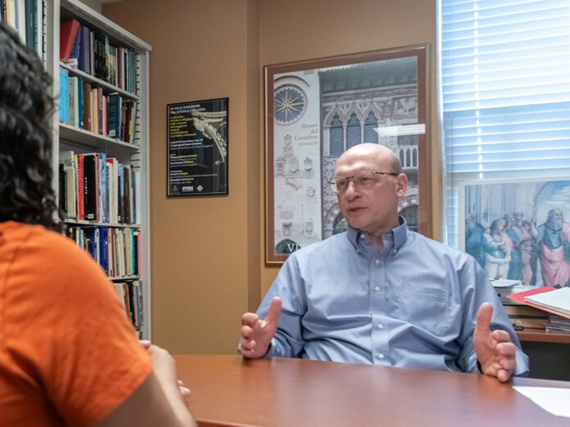 Faculty member sitting behind a desk, talking to a student, with historical images displayed on the wall behind them.