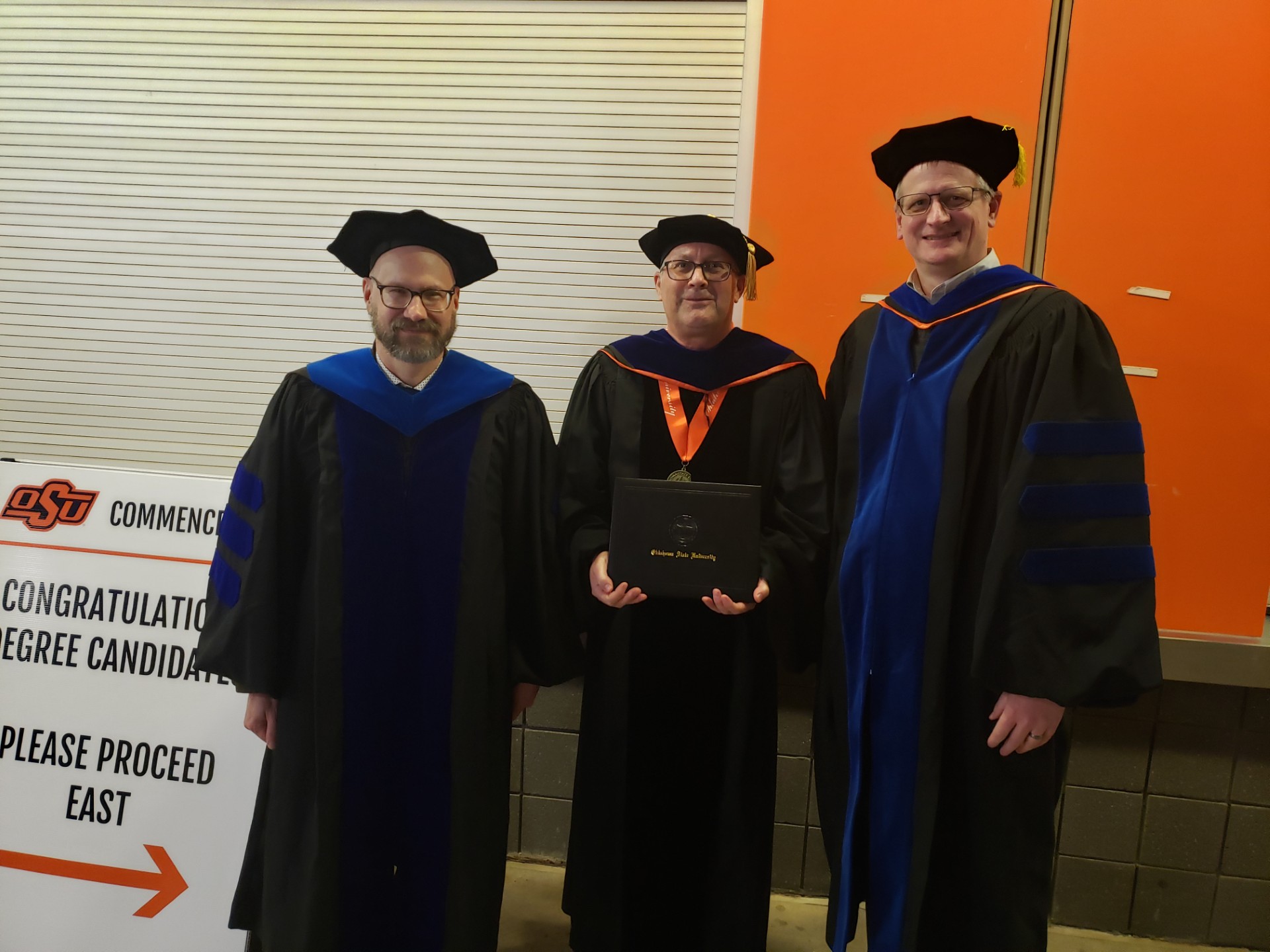 three men stand in graduation gowns with the central man holding a university diploma