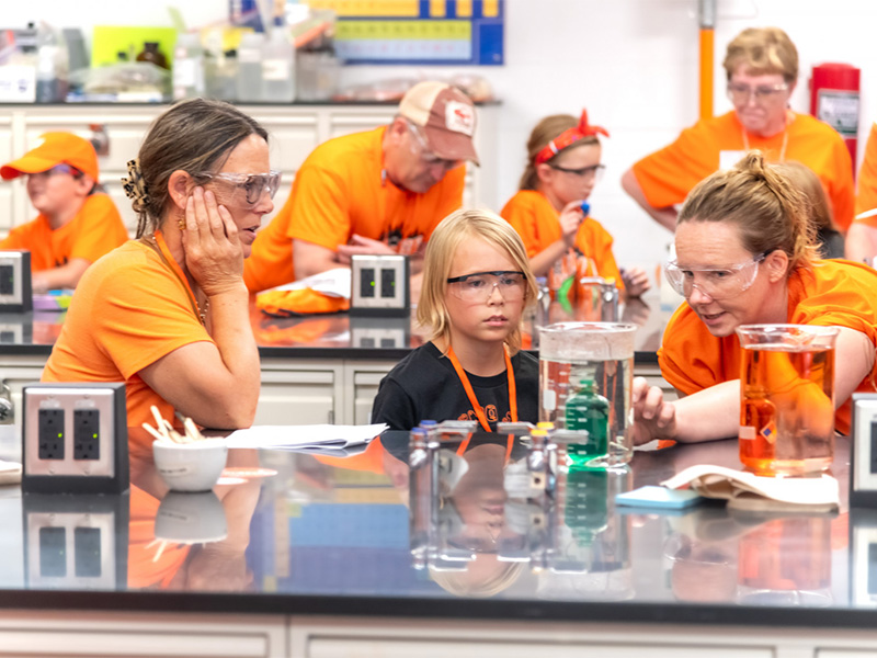 A group of children, parents, and grandparents conducting an experiment in a lab setting, with adults guiding the kids as they work with scientific equipment and materials.