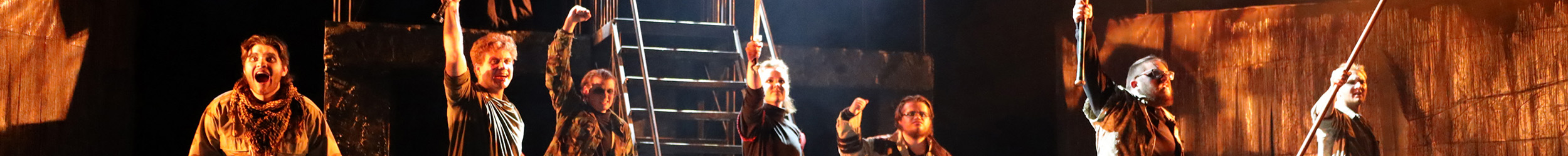 Panoramic stage scene from a dress rehearsal of Peter and the Starcatcher, showing several actors in costume under warm lighting; some hold props like ropes and a sword.